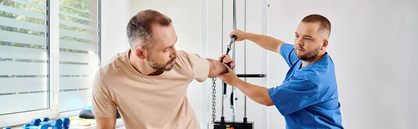 stock image man in sportswear working out on exercise machine near skilled instructor of kinesio center, banner