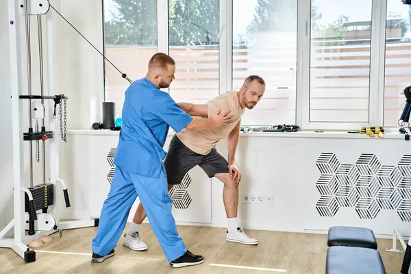 stock image professional instructor in blue uniform helping man during recovery training in kinesiology center