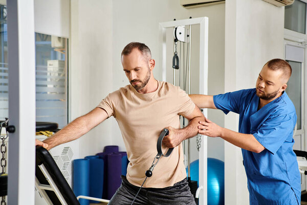 young skilled physician supporting man in sportswear during recovery training on exercise machine
