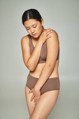sad asian woman in underwear covering body while embracing herself on grey backdrop, body shaming clipart
