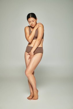 upset asian woman in underwear covering body while embracing herself on grey backdrop, body shaming clipart
