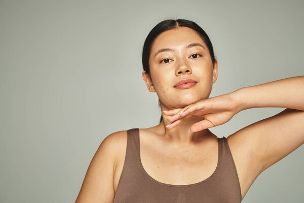 pretty asian woman in brown top touching her face on grey background, skin care and beauty