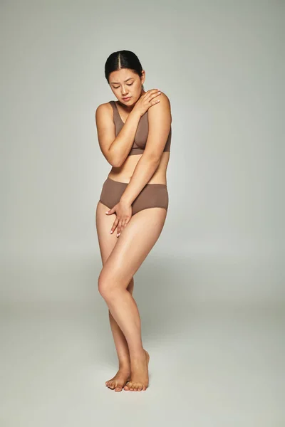 upset asian woman in underwear covering body while embracing herself on grey backdrop, body shaming