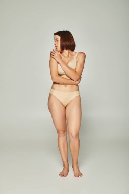 full length of shy woman in beige underwear looking over shoulder on grey background, vulnerability clipart
