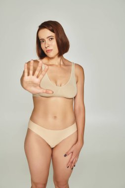 young woman in beige underwear gesturing and showing stop on grey background, body shaming clipart