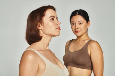 Kısa saçlı, gri arka planda bulanık Asyalı bir kadının yanında poz veren genç bir kadın.