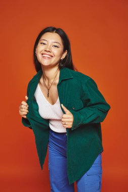 radiant young asian girl posing in vibrant outfit and wearing green jacket on orange background clipart