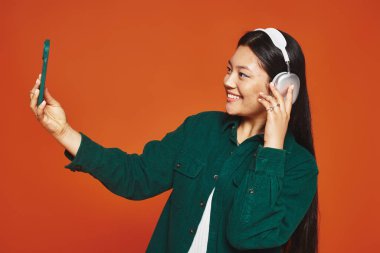 pozitif Asyalı kadın kablosuz kulaklıklarla müzikten hoşlanıyor ve turuncu arka planda selfie çekiyor.