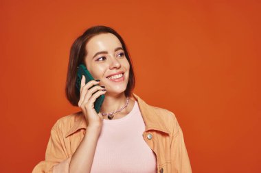 Canlı giyinmiş mutlu genç bir kadın turuncu arka planda akıllı telefondan konuşuyor.