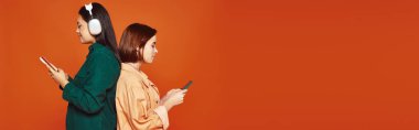Kablosuz kulaklıklı mutlu Asyalı kız turuncu pankarttaki arkadaşının yanında akıllı telefon tutuyor.
