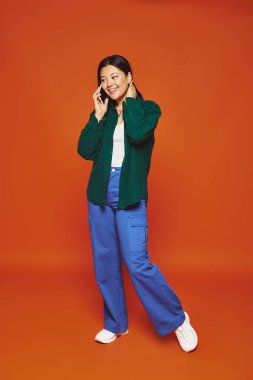 Kumral saçlı mutlu Asyalı kadın akıllı telefondan konuşuyor ve turuncu arka planda gülümsüyor.