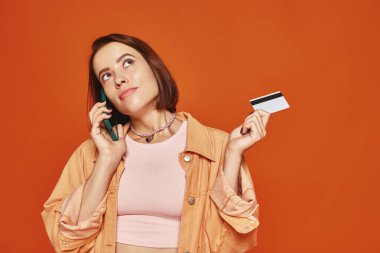 Düşünceli bir kadın akıllı telefondan konuşuyor ve kredi kartını turuncu arka planda tutuyor, online alışveriş yapıyor.