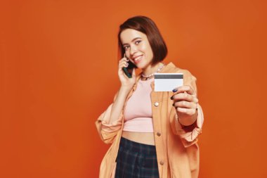 Akıllı telefondan konuşan neşeli bir kadın ve turuncu arka planda kredi kartı tutan, online alışveriş yapan.