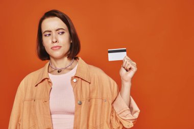 Kısa saçlı düşünceli genç bir kadın kredi kartını turuncu arka planda tutuyor, kişisel finansman.