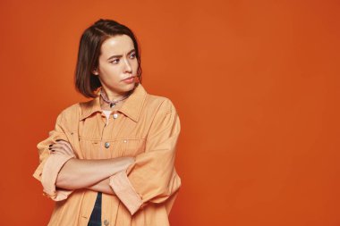thoughtful young woman in casual wear standing with crossed arms on orange background, curious clipart