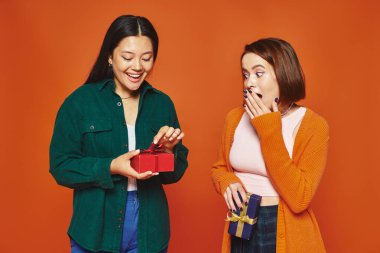 young woman covering mouth and looking at asian friend opening present on orange background clipart