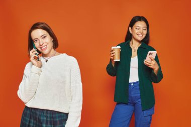 neşeli ve genç çok kültürlü kadınlar akıllı telefonlar kullanıyor ve turuncu arka planda duruyorlar.