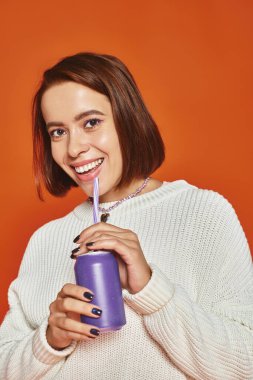 stylish young woman in white knitted sweater sipping soda drink from straw on orange backdrop clipart