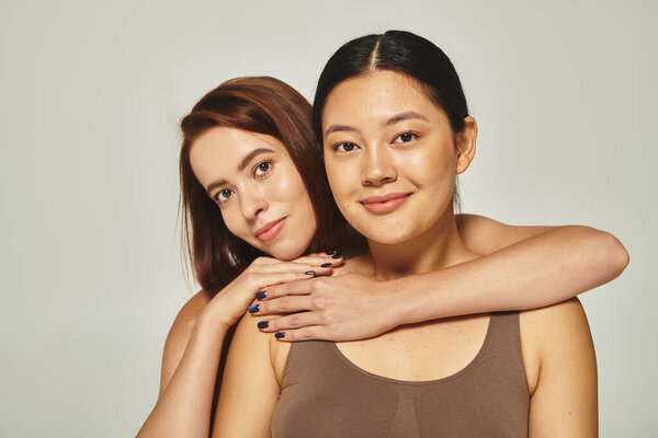 счастливая молодая женщина в нижнем белье обнимает азиатского друга на сером фоне, женская дружба