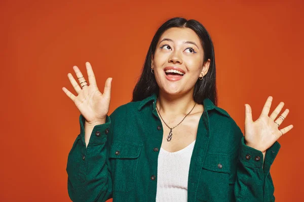 stock image happy asian woman posing in vibrant outfit with green jacket waving hands on orange background