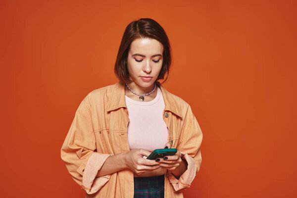 молодая женщина в стильной одежде смс на смартфоне на ярком оранжевом фоне, социальные сети