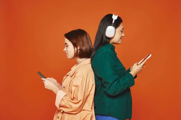 happy asian girl in wireless headphones holding smartphone near her female friend, back to back