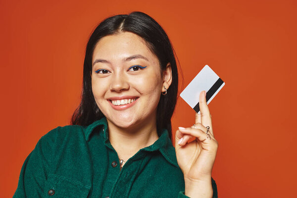 happy young asian woman in green jacket holding credit card on orange background, consumerism