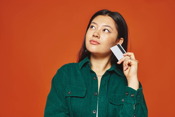 задумчивая молодая азиатка в зеленой куртке с кредиткой на оранжевом фоне, потребительство