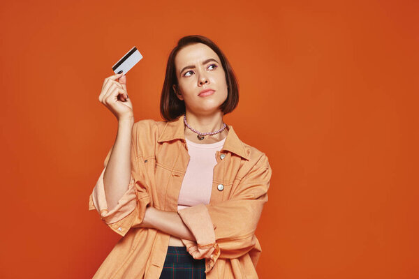 pensive young woman with short hair holding credit card on orange background, personal finance