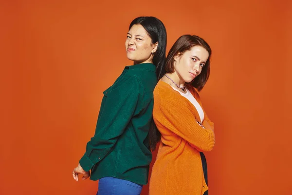 two women in casual clothing standing back to back and looking at camera on orange background