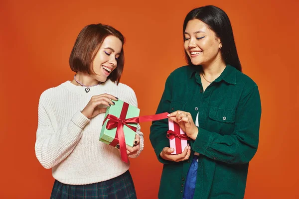 stock image excited young multicultural female friends holding presents on orange background, gift exchange