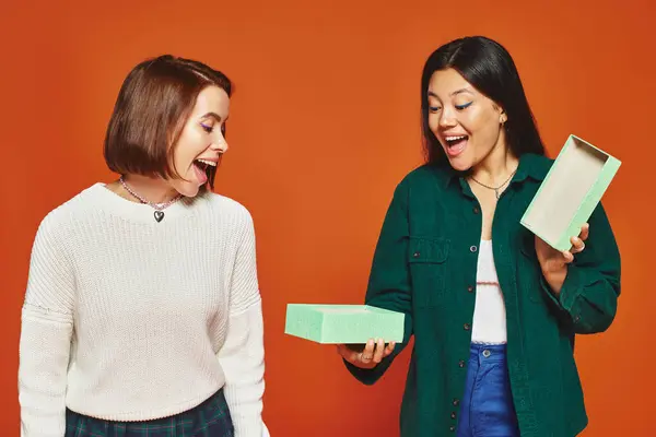 stock image excited asian woman opening present near happy female friend on orange background, gift giving