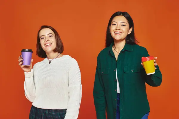 two multicultural female friends holding paper cups with coffee to go on orange background