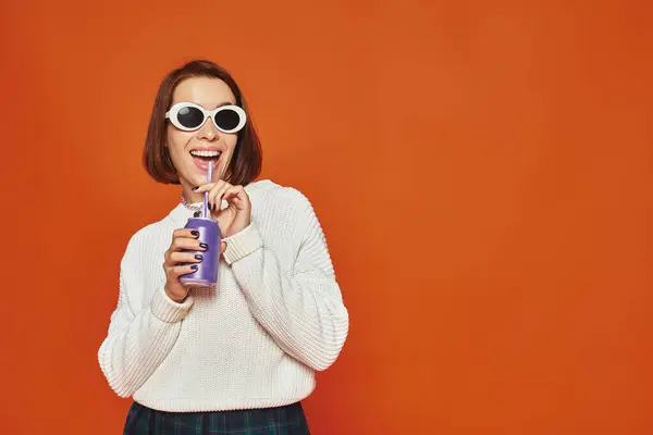 stock image Stylish young woman in white trendy sunglasses sipping soda drink from can on orange backdrop