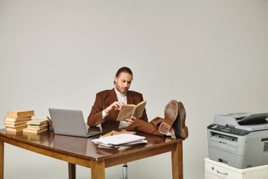 attractive pensive man with beard in brown jacket reading book while in office with legs on table clipart
