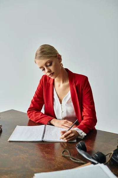 stock image concentrated young woman in red vibrant jacket taking notes while sitting at table in her office