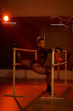 Işıkla aydınlatılan zarif bir kadın, duvardaki parmaklığa yaslanır, esnek bir şekilde.