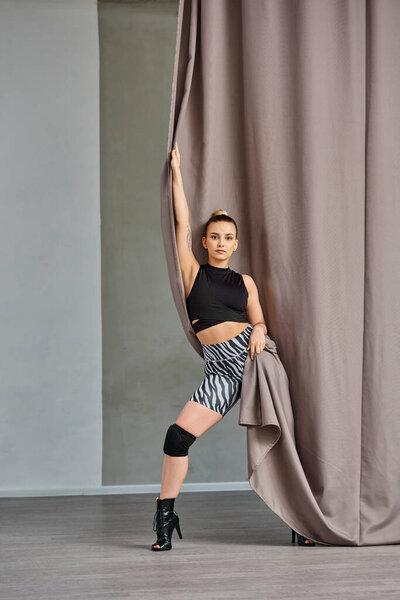 expressive woman in dance clothing and high heels gracefully poses near curtain