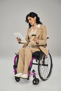 Tekerlekli sandalyede, tablete bakıp elinde kahve tutan, pastel renkli şık giyimli, engelli bir kadın.