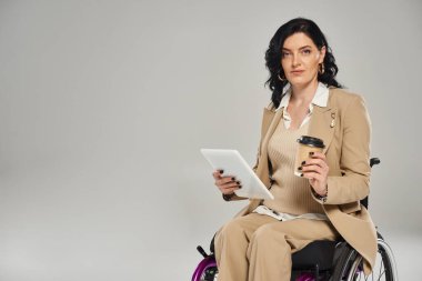Tekerlekli sandalyede kahve ve tabletle kameraya bakan Pastel giysili çekici engelli kadın.