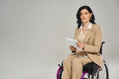 Tekerlekli sandalyedeki çekici, engelli kadın pastel kıyafetler giyiyor, elinde tablet var ve kameraya bakıyor.