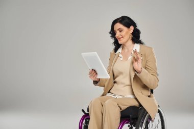 Video çağrısı sırasında tekerlekli sandalyede tablete el sallayan pastel giysili neşeli, engelli bir kadın.