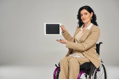 Zarif takım elbiseli çekici bir kadın tekerlekli sandalyede oturuyor ve tabletini kameraya gösteriyor.