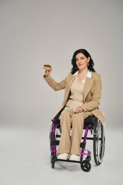 Tekerlekli sandalyede oturan ve kredi kartını kameraya gösteren Pastel takım elbiseli çekici engelli kadın.