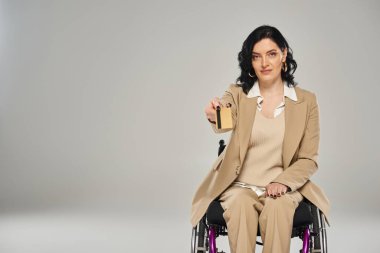 Tekerlekli sandalyedeki güzel, engelli kadın pastel elbise giyiyor. Kameraya kredi kartı gösteriyor.