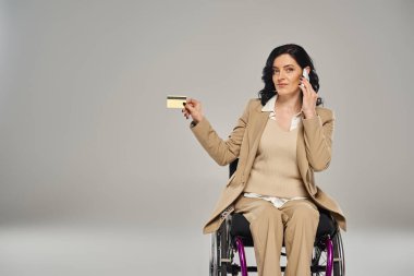 Tekerlekli sandalyedeki pastel kıyafetli kendine güvenen engelli kadın kredi kartı tutuyor ve telefonla konuşuyor.