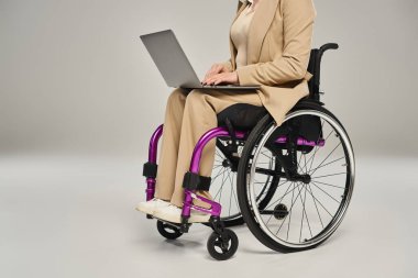 Pastel şık takım elbiseli engelli bir kadın tekerlekli sandalyede oturuyor ve dizüstü bilgisayarla çalışıyor.