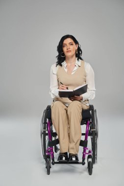 Özürlü çekici bir kadın tekerlekli sandalyede oturmuş elinde not defteriyle kameraya bakıyor.
