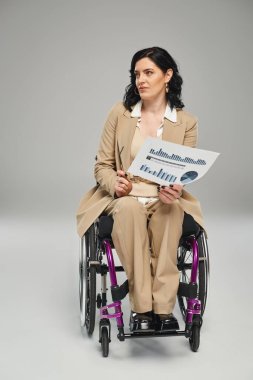 Tekerlekli sandalyedeki pastel kıyafetli çekici engelli kadın elinde grafikler ve gözlüklerle başka tarafa bakıyor.