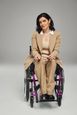 Pastel kıyafetli, tekerlekli sandalyeli, elinde gözlük olan ve kameraya bakan kendine güvenen güzel bir kadın.